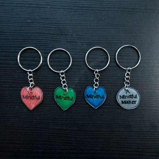 A set of colorful heart-shaped keychains with the words 'Mindful' and 'A Mindful Maker' printed on them, displayed on a dark background.