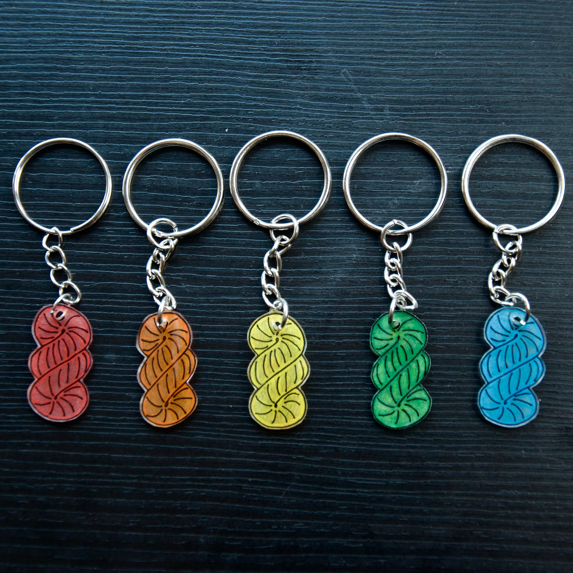 A set of colorful hand-drawn keychains made from recycled plastic, displayed on a dark wooden surface. Each keychain has a silver split loop and chain attachment.