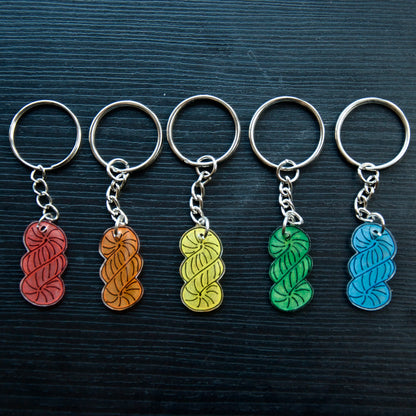 A set of colorful hand-drawn keychains made from recycled plastic, displayed on a dark wooden surface. Each keychain has a silver split loop and chain attachment.
