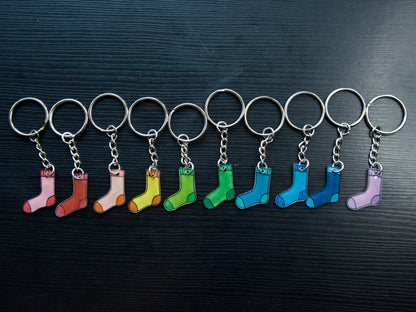 A collection of hand-drawn keychains in various colors shaped like socks, attached to silver split loops with chain attachments, displayed on a dark surface.