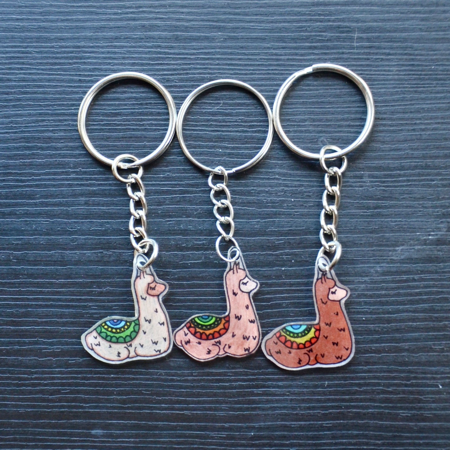 A collection of hand-drawn keychains with various cute designs, attached to silver split loops with chains. The keychains are made from recycled plastic and are in different colors, featuring simple, minimalistic alpaca designs.