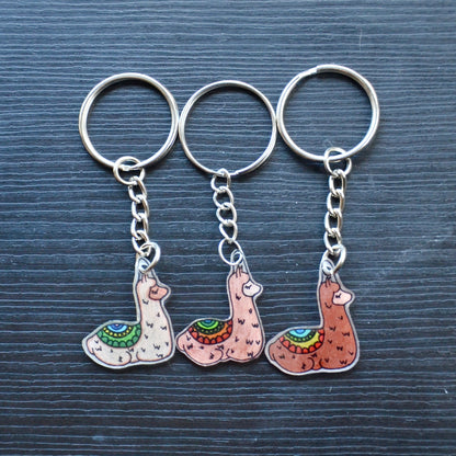 A collection of hand-drawn keychains with various cute designs, attached to silver split loops with chains. The keychains are made from recycled plastic and are in different colors, featuring simple, minimalistic alpaca designs.