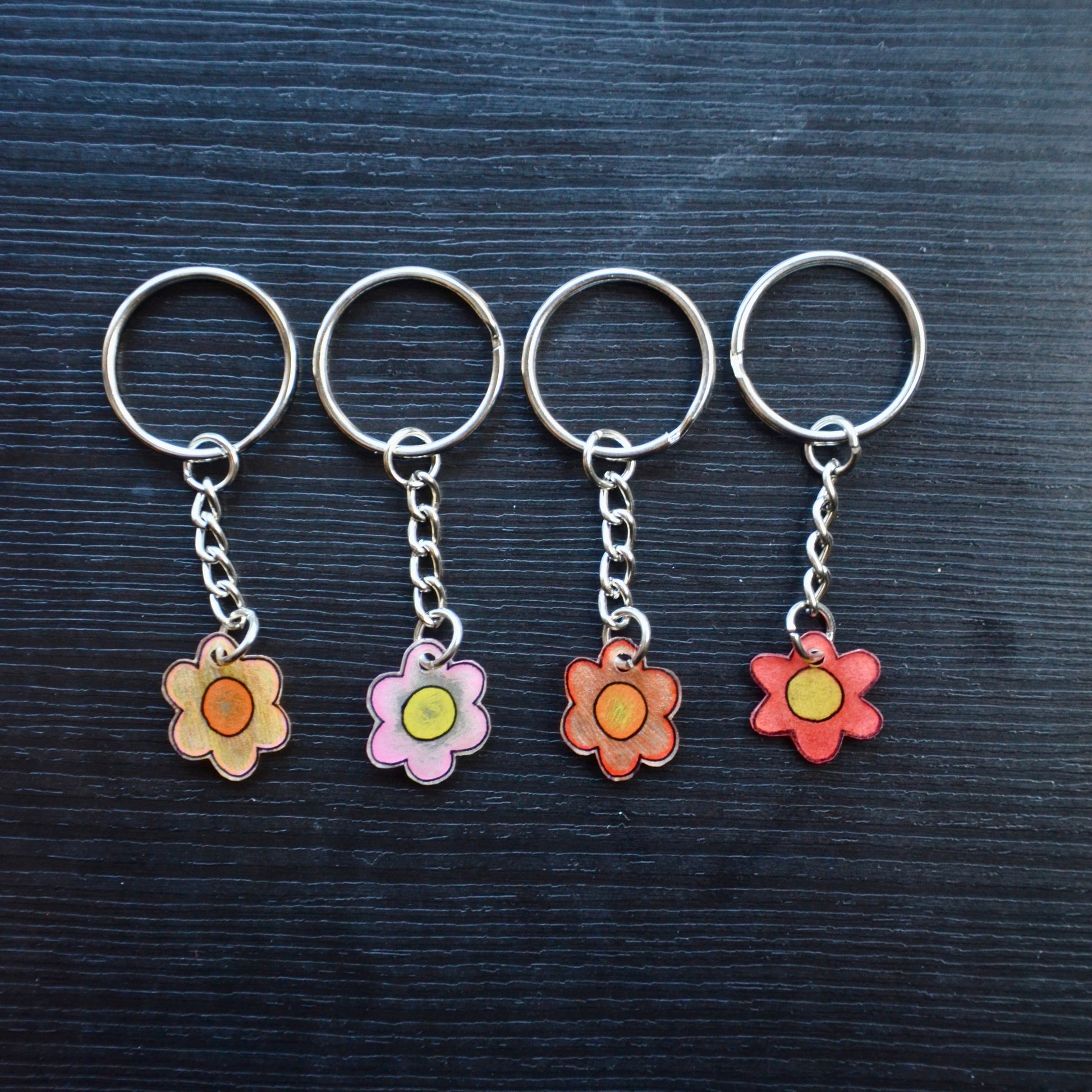 Four keychains with flower designs, one with a pink gradient and the other with a yellow-orange gradient, attached to silver split loops with chains on a dark background.