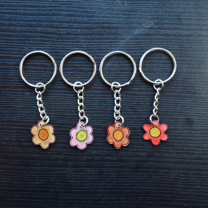 Four keychains with flower designs, one with a pink gradient and the other with a yellow-orange gradient, attached to silver split loops with chains on a dark background.