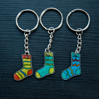 A collection of hand-drawn keychains in various colors shaped like socks, attached to silver split loops with chain attachments, displayed on a dark surface.