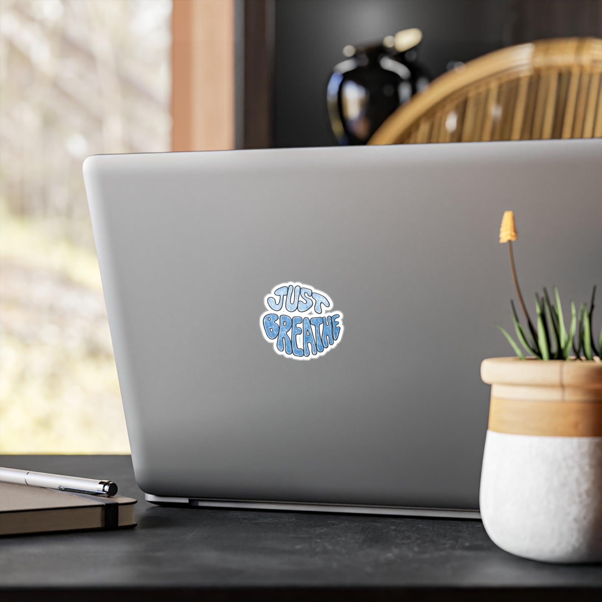 Laptop with a sticker of "Just Breathe" on a desk with a plant and pen in the foreground