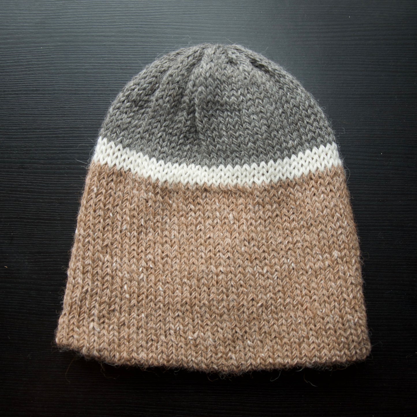 A reversible hiking beanie with one side beige and the other side gray, made from alpaca yarn, displayed on a dark background.