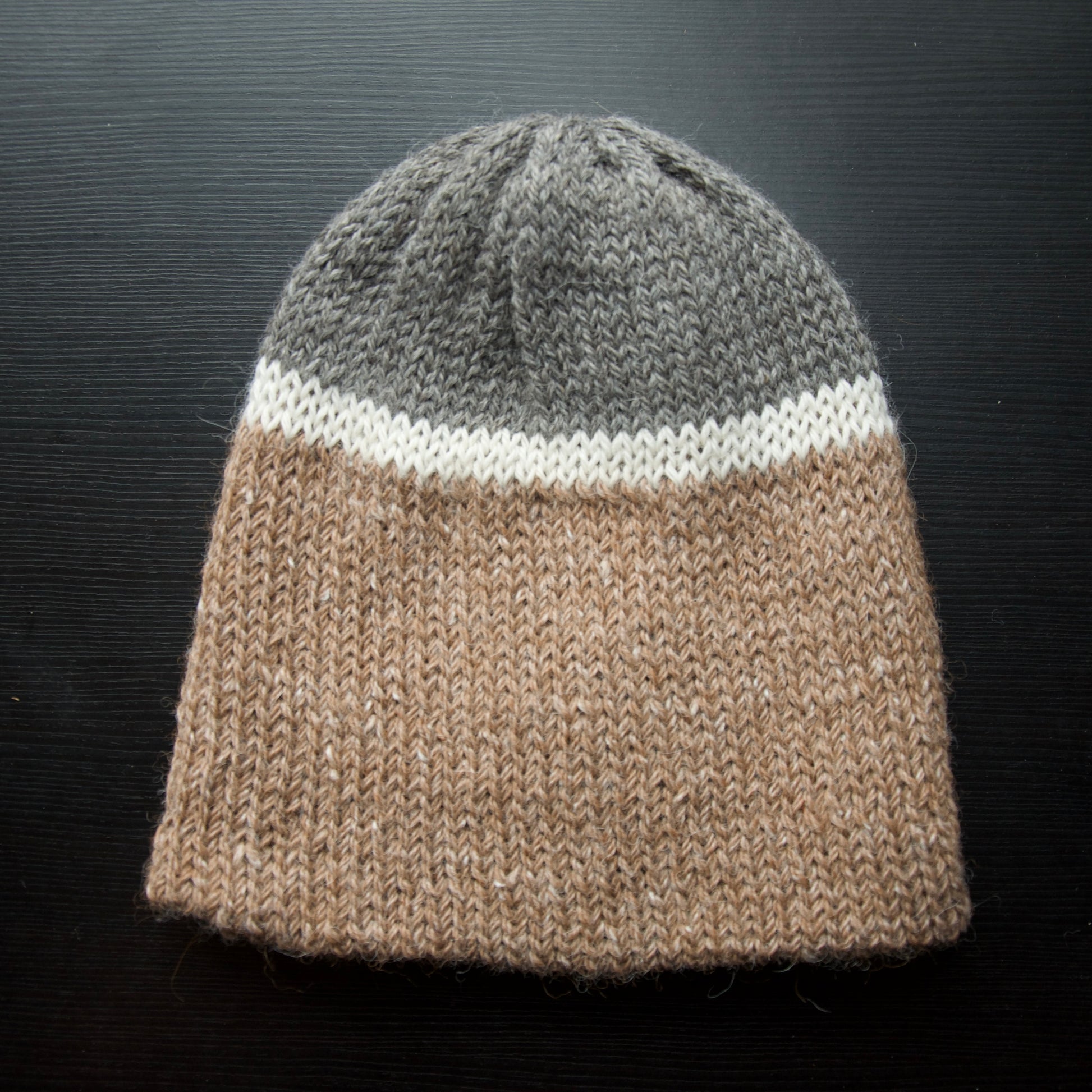 A reversible hiking beanie with one side beige and the other side gray, made from alpaca yarn, displayed on a dark background.