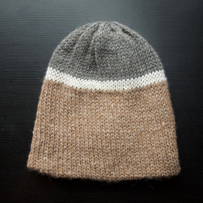 A reversible hiking beanie with one side beige and the other side gray, made from alpaca yarn, displayed on a dark background.