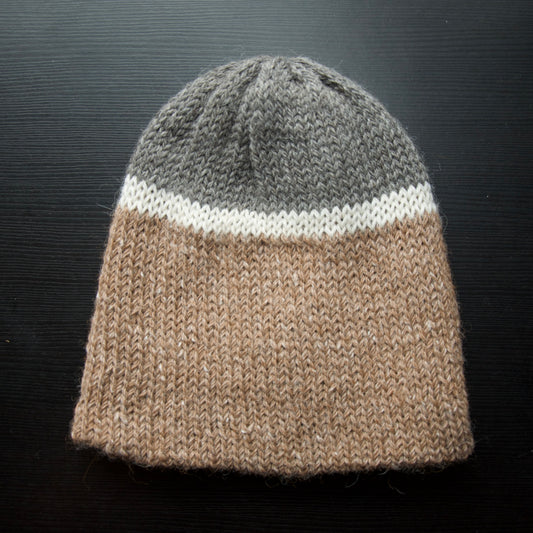 A reversible hiking beanie with one side beige and the other side gray, made from alpaca yarn, displayed on a dark background.
