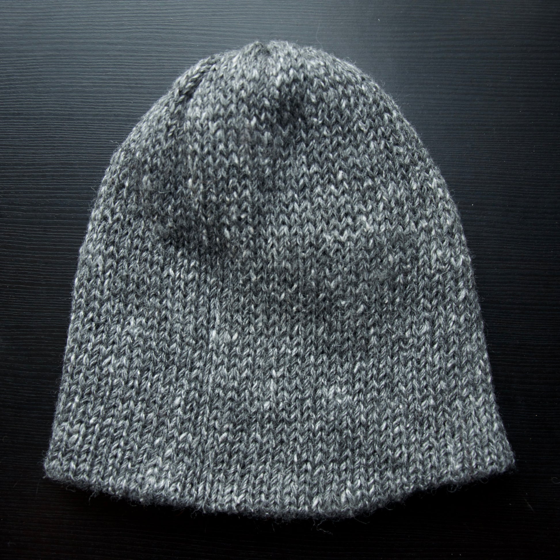 A reversible hiking beanie with one side beige and the other side gray, made from alpaca yarn, displayed on a dark background.