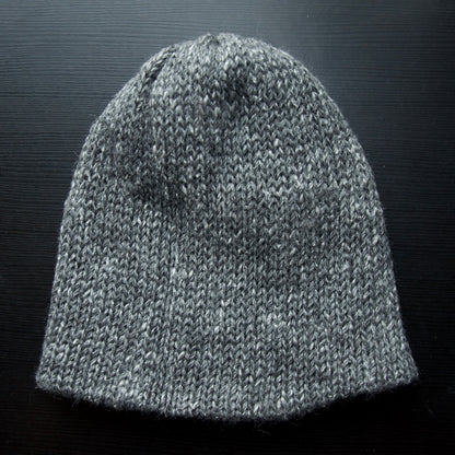 A reversible hiking beanie with one side beige and the other side gray, made from alpaca yarn, displayed on a dark background.