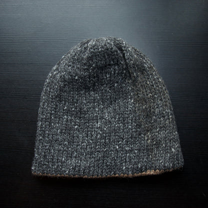A reversible hiking beanie with one side beige and the other side gray, made from alpaca yarn, displayed on a dark background.