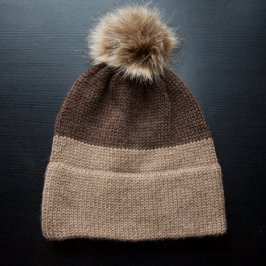 A knitted hat with a beige and brown color block design, featuring a removable pom-pom on top.
