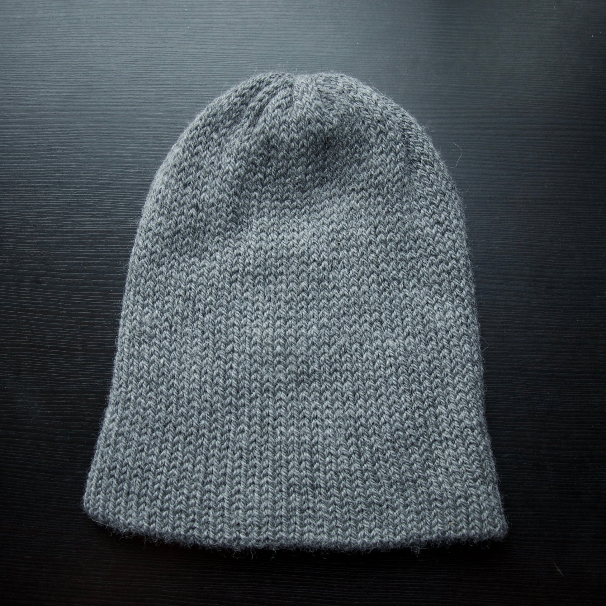 A reversible hiking beanie with one side beige and the other side gray, made from alpaca yarn, displayed on a dark background.