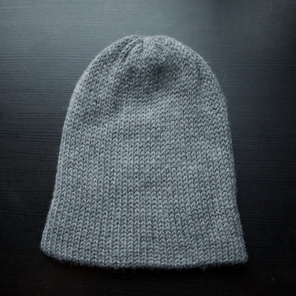 A reversible hiking beanie with one side beige and the other side gray, made from alpaca yarn, displayed on a dark background.