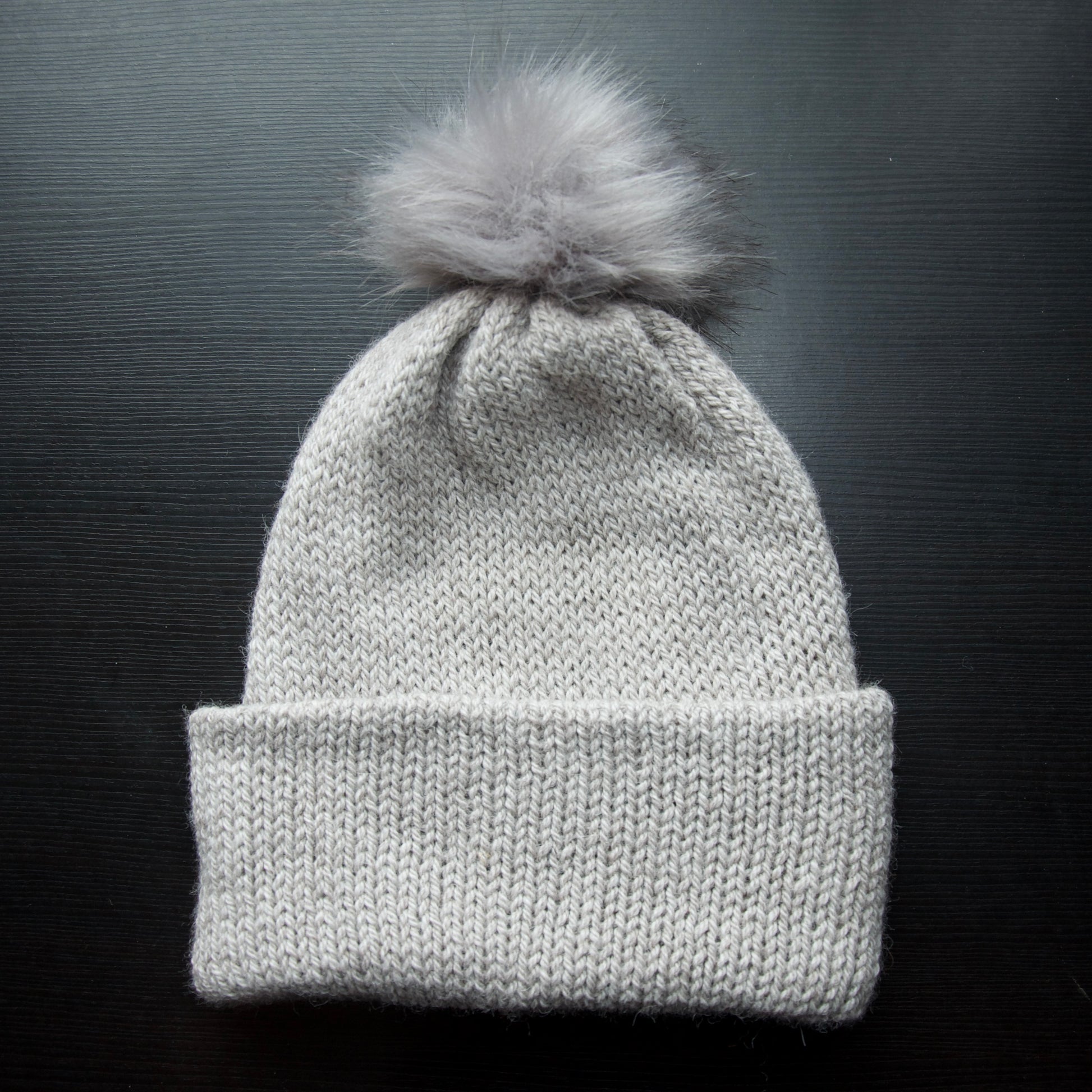 A gray beanie with a removable pom pom on a dark background.
