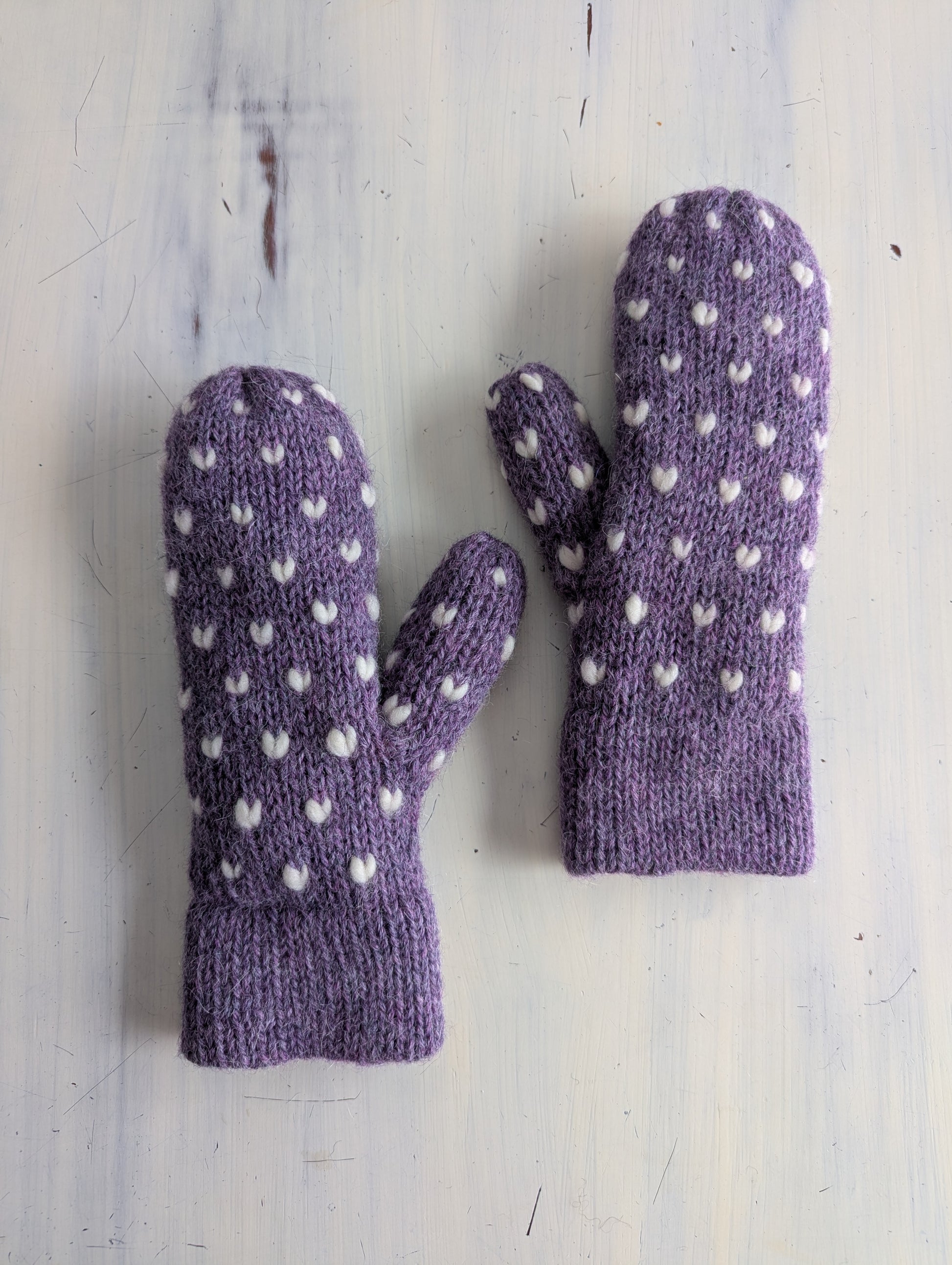 Pair of purple thrun mittens with white hearts on a light wooden surface