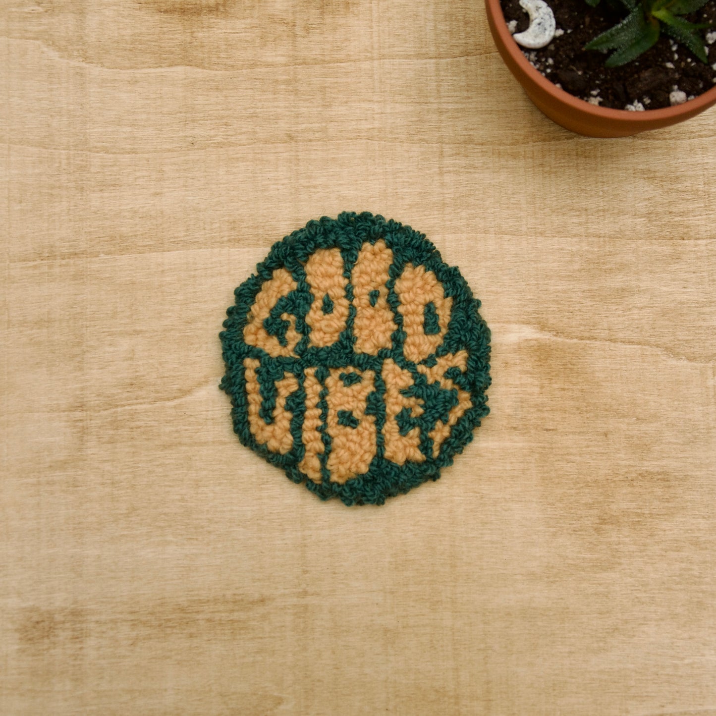 A hand-stitched, circular coaster with the words 'GOOD VIBES' written on it, placed on a beige surface with a potted plant in the background.