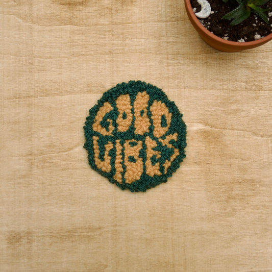 A hand-stitched, circular coaster with the words 'GOOD VIBES' written on it, placed on a beige surface with a potted plant in the background.