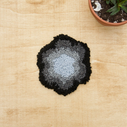 A hand-stitched, punch needle coaster made of various fibers, displayed on a beige background.