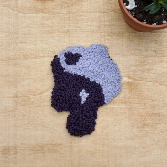 Hand-stitched punch needle coaster made of various fibers, displayed on a beige background with a small potted plant beside it.