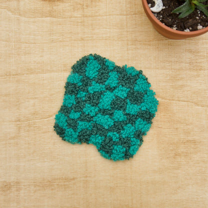 Hand-stitched punch needle coaster made of various fibers, displayed on a beige background with a small potted plant beside it.